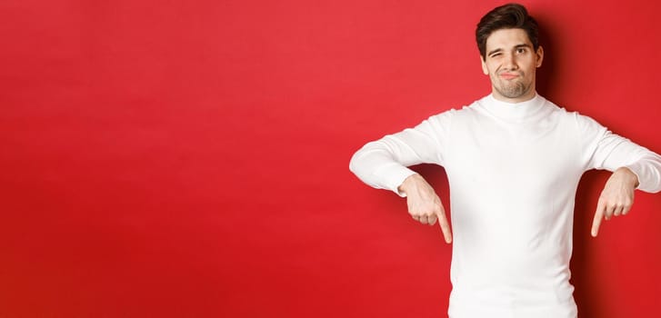 Portrait of attractive, skeptical young man in white sweater, grimacing and pointing fingers down, disagree and dislike something, standing over red background.