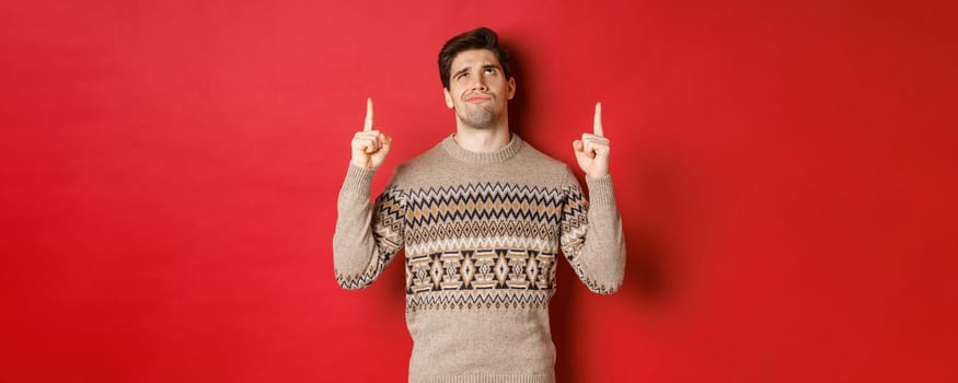 Image of disappointed and skeptical handsome man, wearing christmas sweater, smirk and frowning as looking at something bad, pointing fingers up, standing over red background.