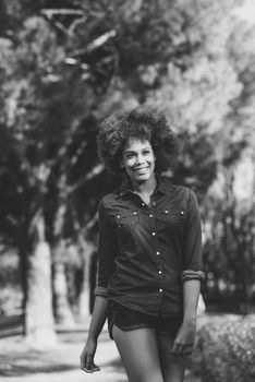 Young black woman with afro hairstyle walking in urban park. Mixed woman wearing blue shirt and shorts. Female smiling.