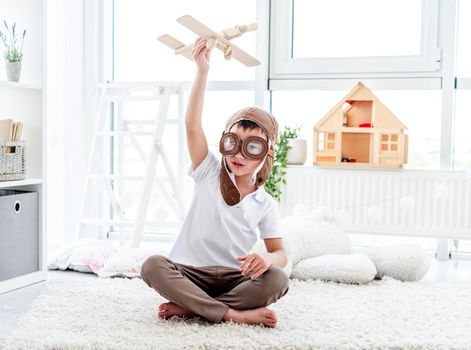 Happy little boy in pilot hat playing with wooden plane in children's room