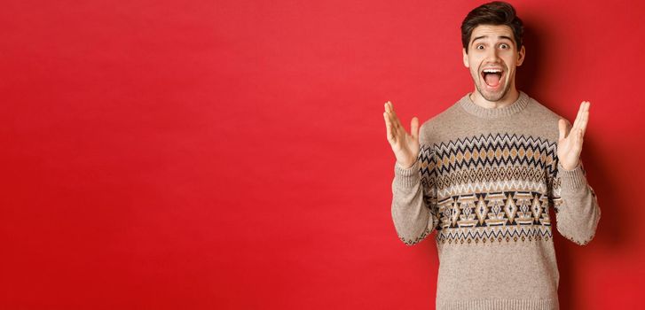 Portrait of surprised and happy handsome man, wearing christmas sweater, looking amazed, celebrating new year, standing over red background.