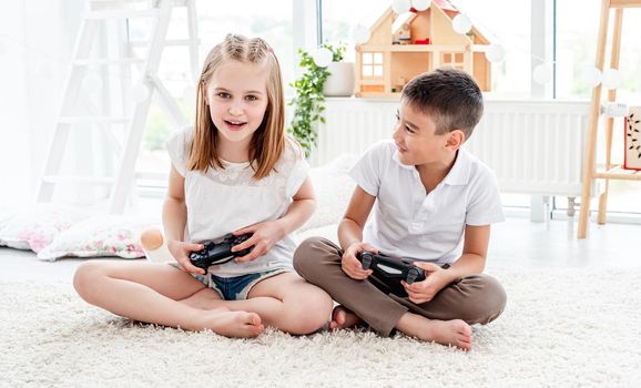 Playful kids with joysticks for video gaming sitting on floor in light room