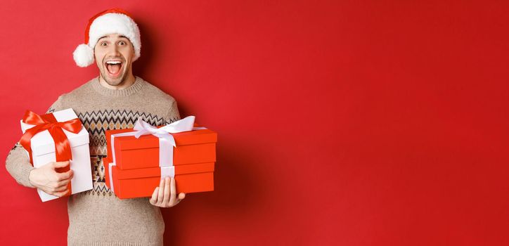 Concept of winter holidays, new year and celebration. Image of amazed and happy man in sweater and santa hat, holding christmas gifts and shouting for joy, red background.