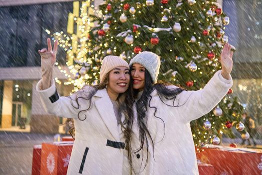 Two girlfriends asian girls in white coats are having fun and hugging in front of Christmas Tree, new year mood