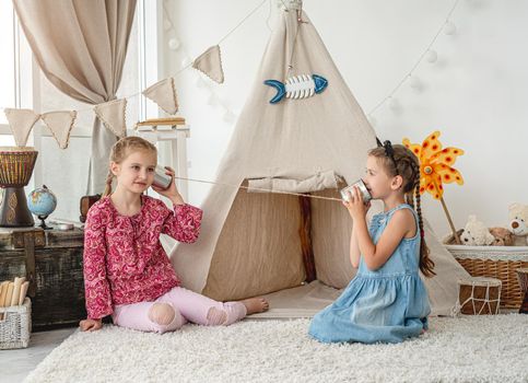 Little sisters playing with homemade telephone made of can and thread in room with wigwam