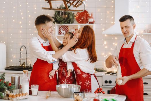 A happy family is standing in the Christmas kitchen and smearing each other with flour.