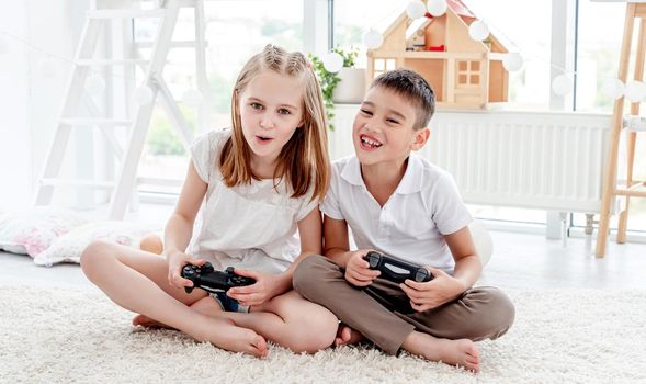 Cheerful little boy with hands up and cute little girl playing video game sitting in kids room