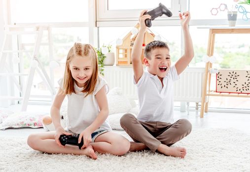 Cheerful little boy with hands up and cute little girl playing video game sitting in kids room