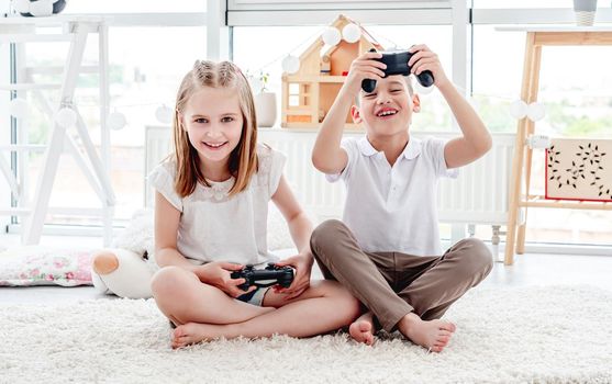 Playful kids with joysticks for video gaming sitting on floor in light room