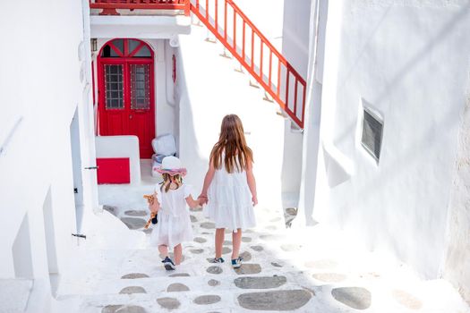 Two little girls in white dresses holding hands white walking down traditional narrow Greek streets ar sunny day