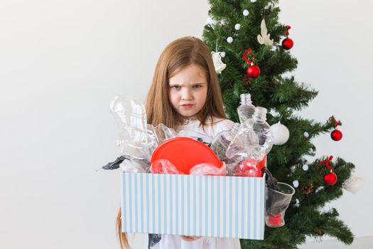 Shocked little child girl looks with opened eyes and worried expression, holding box with various plastic wastes over christmas tree background