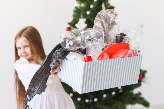 Shocked little child girl looks with opened eyes and worried expression, holding box with various plastic wastes over christmas tree background