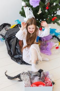 Shocked little child girl looks with opened eyes and worried expression, holding box with various plastic wastes over christmas tree background