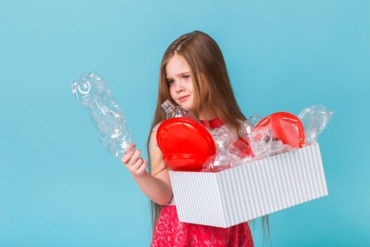 Shocked little child girl looks with opened eyes and worried expression, holding box with various plastic wastes on blue background