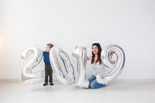 New year, celebration and holidays concept - mother and son sitting near sign 2019 made of silver balloons for new year in white room background.