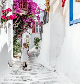 Little child girl in white dress walking the narrow alley with beautiful pink flowers between whitewashed buildings, Greece