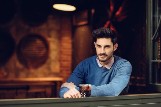 Young man with blue sweater with lost look in a modern pub. Thoughtful guy with modern hairstyle near a window drinking a soda.