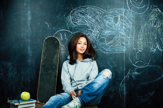 young cute teenage girl in classroom at blackboard seating on table smiling, modern hipster concept closeup