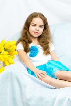 A beautiful little girl with long blond, flowing hair sits on the couch next to a large bouquet of yellow flowers, tulips.
