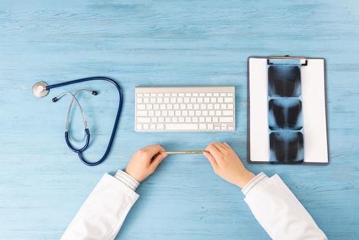 Top view of doctor hands holding pen. X-Ray radiography examination in hospital. Physician sitting at blue wooden desk. Examination and consultation in clinic. Workplace with stethoscope and keyboard.