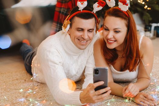 a happy married couple is lying on the floor at home near the Christmas tree and taking pictures of themselves.