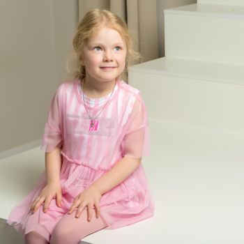 A charming little girl in a smart dress sits on a white staircase. The concept of happiness, childhood.