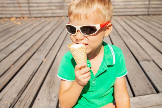 Adorable blond haired little boy in fashionable sunglasses biting yummy vanilla ice cream cone while spending summer day in park