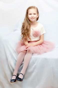 Caucasian little girl with long blond hair, in a beautiful pink skirt and white T-shirt and a pink bow on her chest.Sits on a blue sofa.