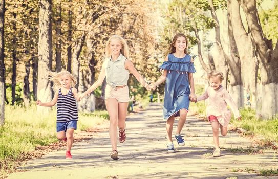 Front view of little smiling blond girls running on the sunshine autumn alley by holding hands