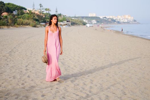 Portrait of a beautiful woman with long pink dress and sun hat on a tropical beach