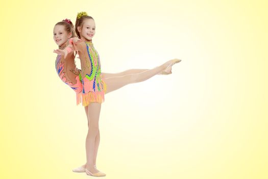 Two adorable little twin girls, gymnastics in the sports school. Girls beautiful gymnastic leotards. They do the splits.On a yellow gradient background.