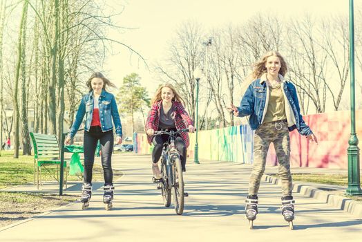 Group of three teen girls in clourful trendy clothes having fun while roller skating and riding a bicycle at park