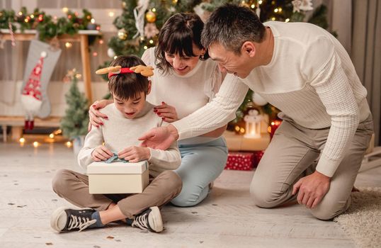 Smiling little boy unboxing christmas present while sitting beside happy parents in festive room