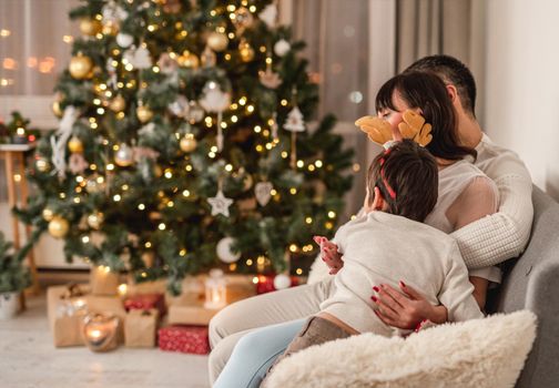 Happy family sitting sideways on sofa next to illuminated christmas tree at home