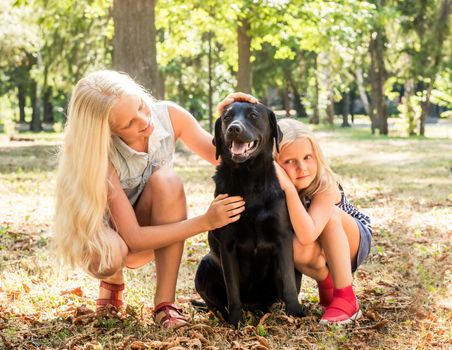 Pretty little blond girls sit hugging a black retriever in a sunshine autumn park