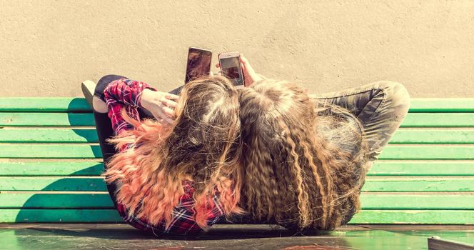 Top shot of two girls using mobile phones while sitting on the bench and talking