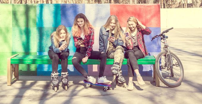 Trendy young girls laughing while using mobile phones in a skateboard park. Four pretty girl friends sitting on the bench with skateboards, rollers and bicycle