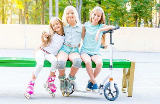 Little smiling blond girls sitting hugging on the green bench with scooter and rollerskates put on