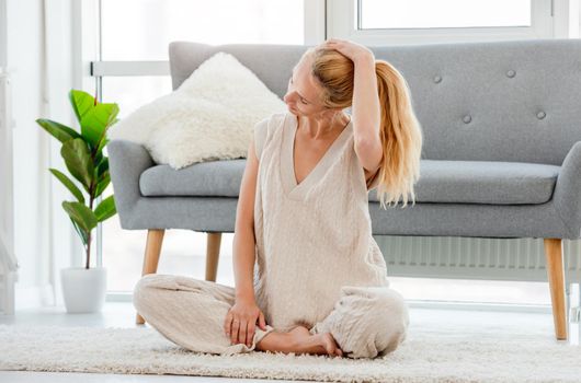 Blond girl sitting on the floor and stretch her neck during morning yoga workout at home