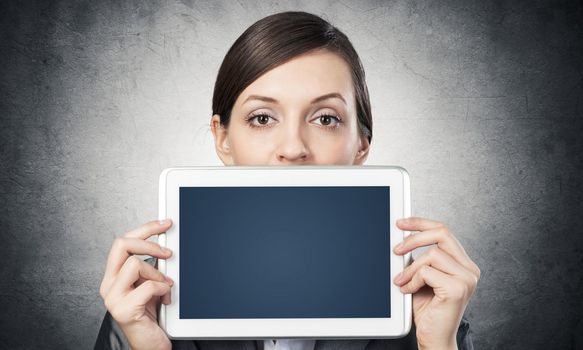 Businesswoman holding tablet computer with blank screen. Beautiful woman in business suit show tablet PC near her face. Corporate businessperson on grey wall background. Digital technology layout.