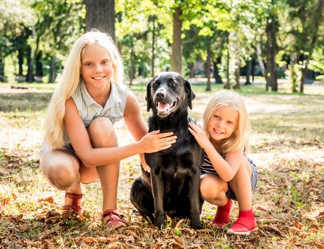 Pretty smiling blond girls sit hugging a black retriever in a sunshine autumn park