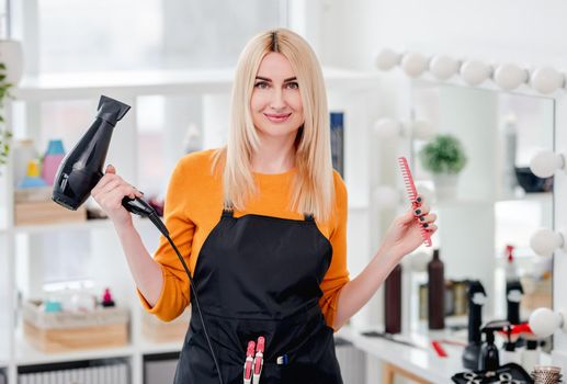 Beautiful blond woman hairdresser looking at camera and smiling with hairdryer in her hand