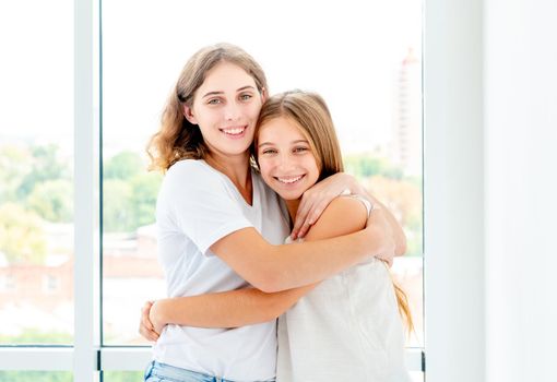 Sisters hug happily in light room near window