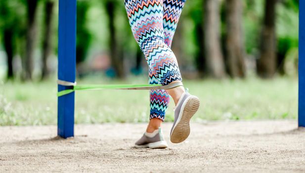 Sport girl with elastic rubber band doing leg workout at stadium. Young woman exercising with fitness equipment outdoors