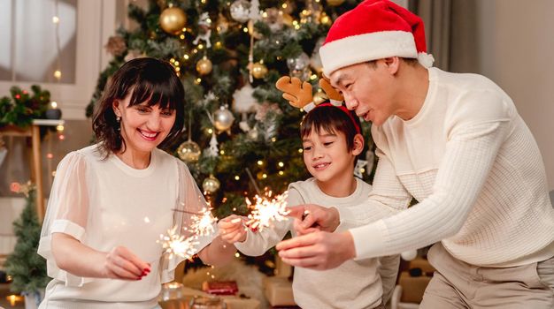Happy family in santa hats with sparklers sitting under decorated christmas tree at home
