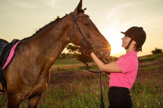 Beautiful smiling girl jockey stand next to her brown horse wearing special uniform on a sky and green field background on a sunset. Equitation sport competition and activity.