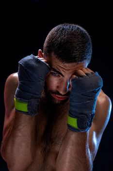 Determined young man with athletic body wearing training bandages posing in defensive position, holding pumped fists in front of his face and staring at camera on black background. Strength and motivation. Studio shot