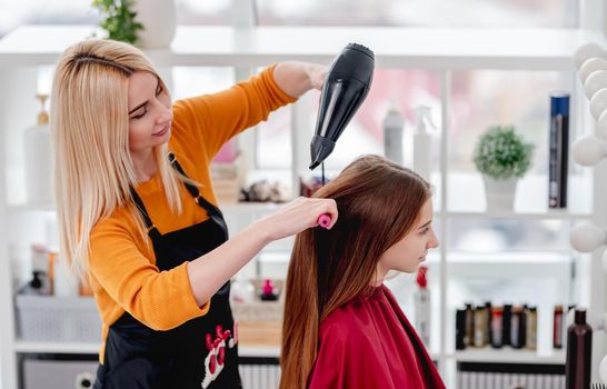 Blond woman hairdresser dries long hair of young girl client with hairdryer in salon