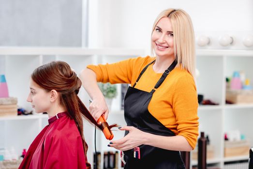 Blond woman hairdresser winds curls for beautiful young model hairstyle with a hair iron and smiling