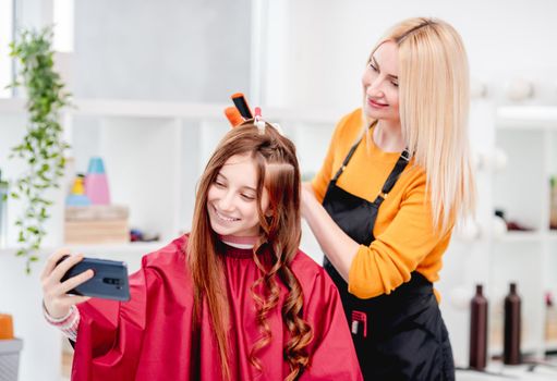 Beautiful young girl making selfie with hairdresser during hairstyle process. Curls hairdo for long hair